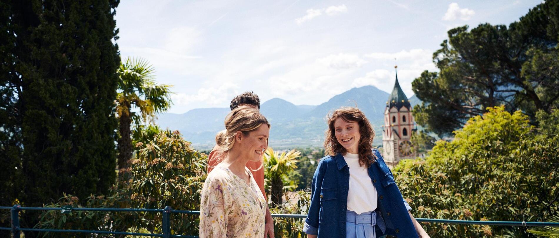 Zwei Frauen und ein Mann spazieren im Frühling entlang einer Promenade in Meran