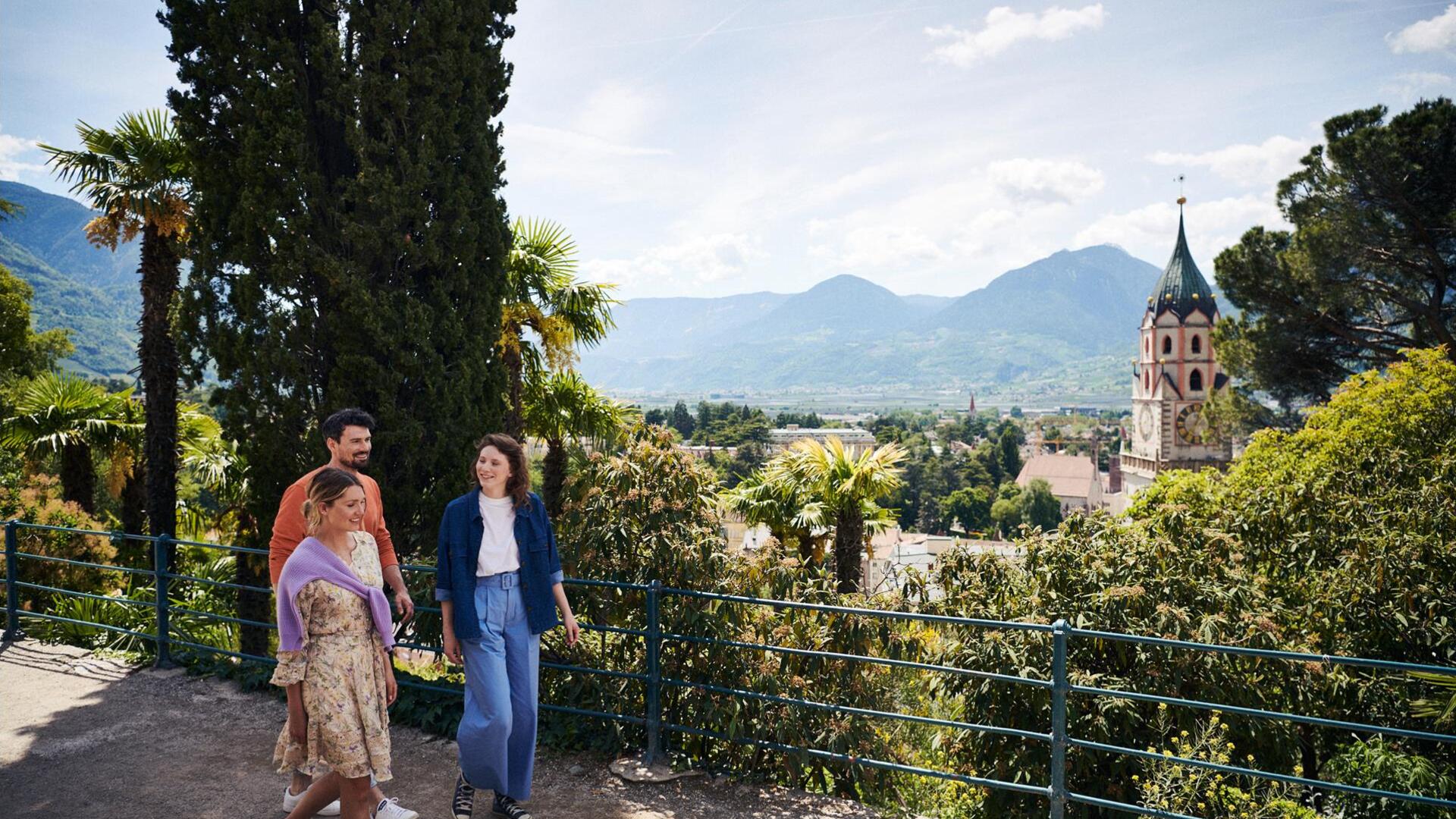 Zwei Frauen und ein Mann spazieren im Frühling entlang einer Promenade in Meran